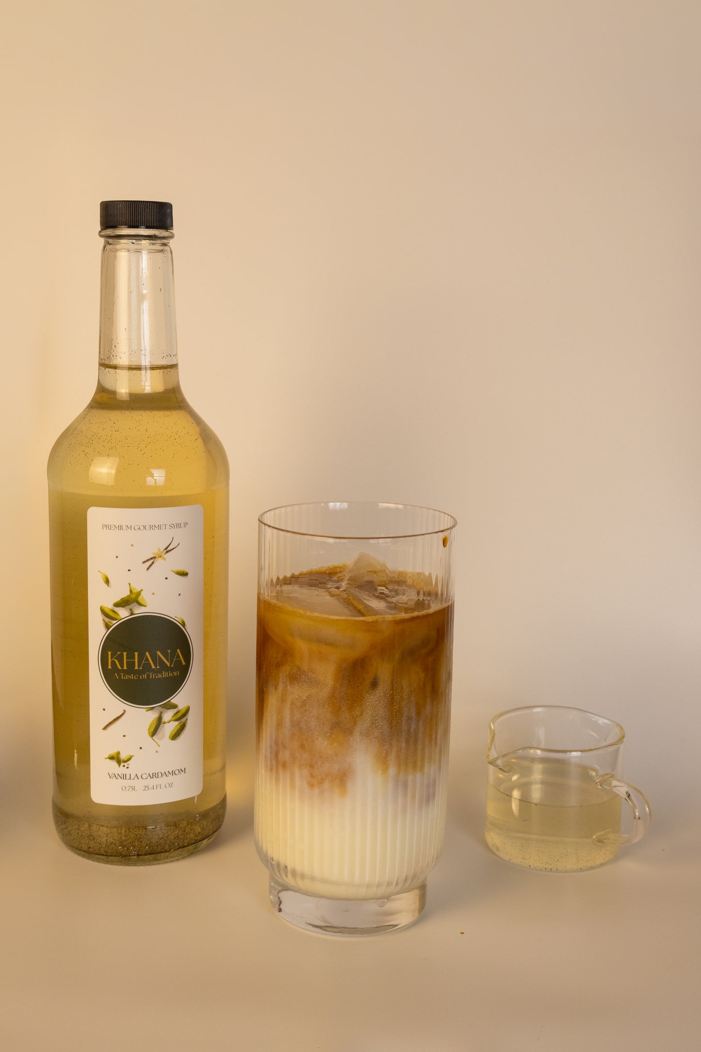 Khana Vanilla Cardamom Syrup 750mL bottle next to a glass of coffee latte and serving of syrup
