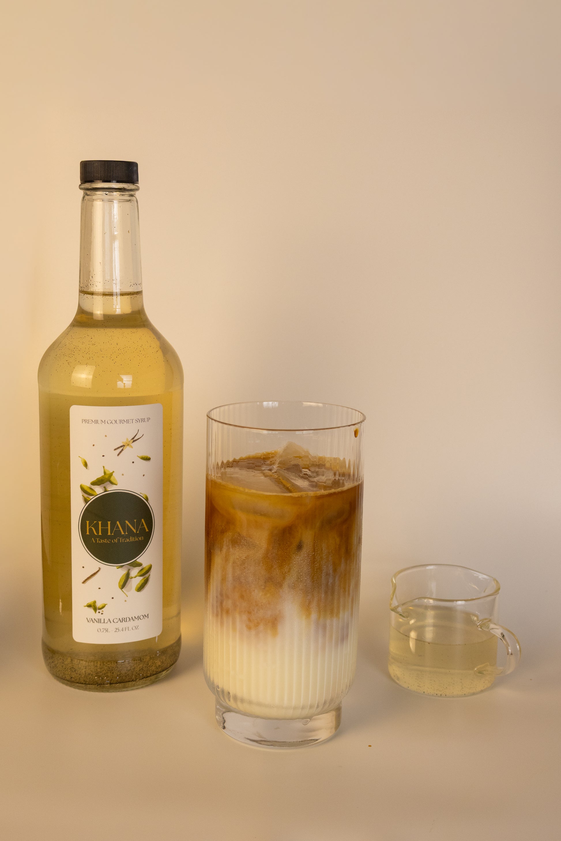 Khana Vanilla Cardamom Syrup 750mL bottle next to a glass of coffee latte and serving of syrup